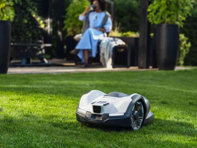 Husqvarna Automower 405VE Nera fährt auf dem Rasen, Frau sitzt im Hintergrund