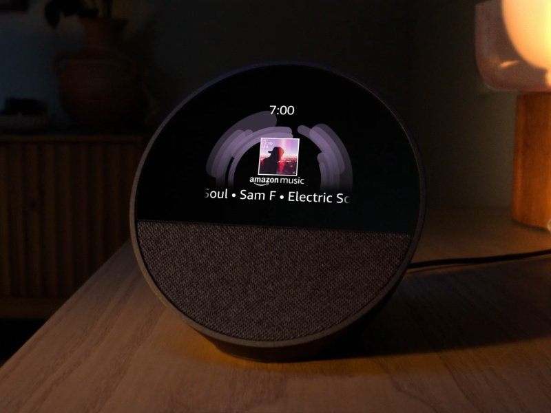 Dunkelgraues Amazon Alexa Smart Display auf einem Holztisch in einem schummrigen Raum, zeigt violette Audiowiedergabe und Albumcover von Amazon Music.