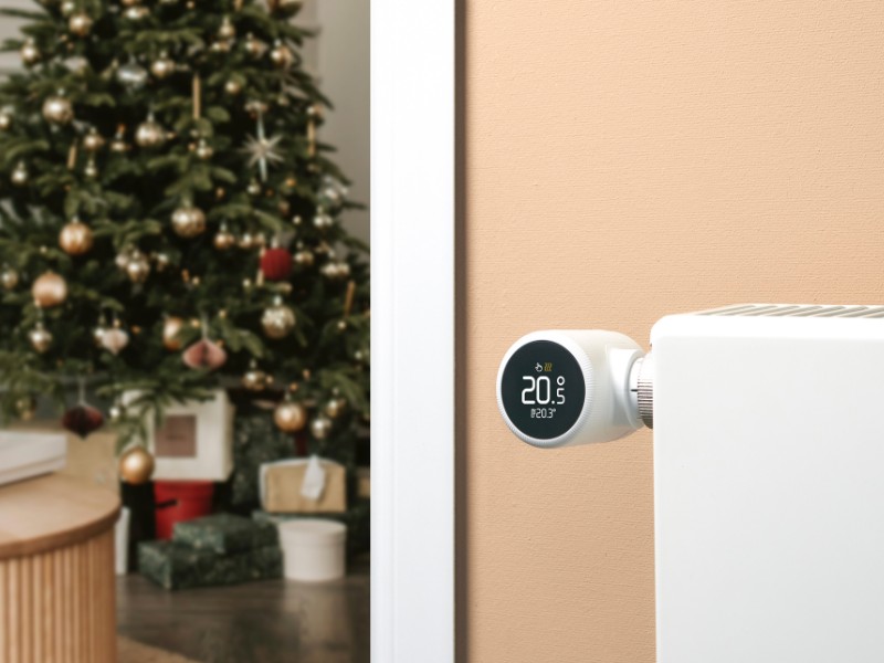 Smartes tado° Thermostat an einem Heizkörper vor einer beigen Wand, eingestellt auf 20,5 Grad, mit einem verschwommenen Weihnachtsbaum im Hintergrund. Energie sparen an Weihnachten.