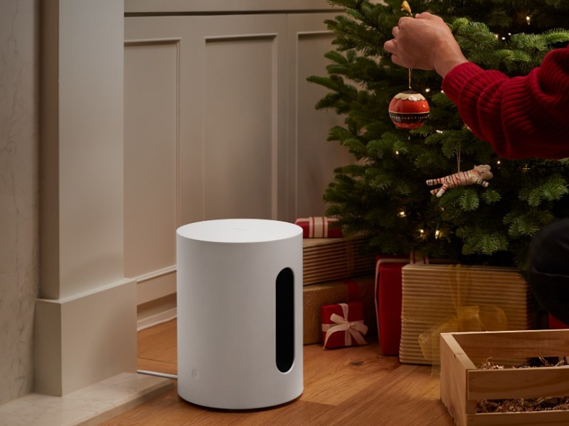 Mann in rotem Pullover schmückt einen Weihnachtsbaum. Ein weißer Sonos Sub Mini steht auf dem warmen Holzboden, umgeben von gold-roten Weihanchtsgeschenken