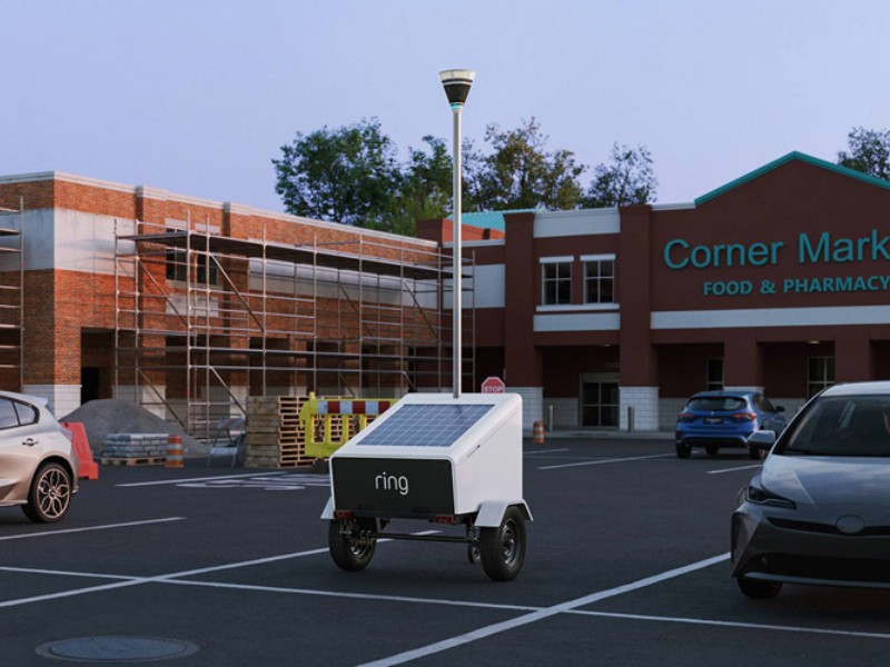 Weiße mobile Ring Elite-Überwachungseinheit mit Solarpanel und hohem Kameramast (CES 2026) auf einem Parkplatz, links ein im Bau befindliches Backsteingebäude und rechts der rotbraune Corner Market.