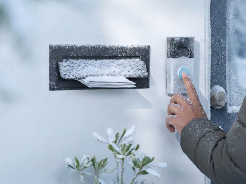Eine Hand in einem grünen Wintermantel drückt den blau beleuchteten Knopf einer schneebedeckten Video-Türklingel (Ring) an einer weißen Tür – ein Hinweis auf den Akku der smarten Türklingel bei Kälte.