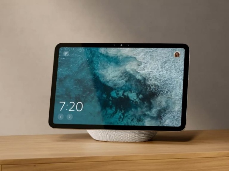 Smart Display mit weißem Textilfuß auf einem hellen Holztisch. Der Bildschirm zeigt die Uhrzeit 7:20 vor einem dynamischen, türkisblauen Meereshintergrund, ideal für die Alexa Tagesplanung.