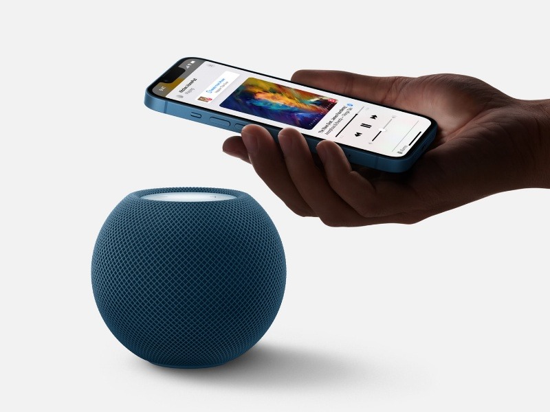 Eine Hand hält ein hellblaues iPhone über einen mitternachtsblauen HomePod mini mit Netzstruktur und einer leuchtend weißen Oberseite. Auf dem iPhone-Bildschirm wird der Text „KitchenHomePod playing“ mit aktiver Musiksteuerung und Albumcovern angezeigt.