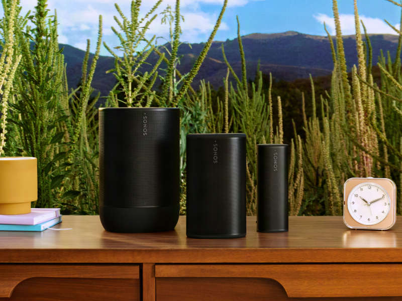 Sonos Move 2, Play und Roam 2 nebeneinander auf einer Kommode mit Uhr, Büchern und Lampe, Natur im Hintergrund