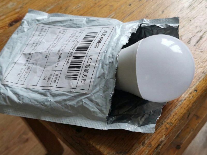 Eine große weiße LED-Lampe ragt aus einem zerrissenen, grauen AliExpress-Versandbeutel mit chinesischen Etiketten heraus, der auf einem abgenutzten Holztisch liegt.