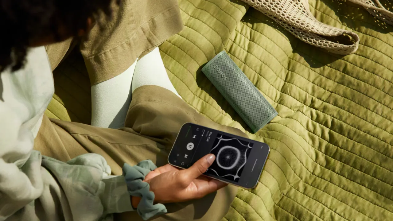 Eine Person steuert die Musik per Smartphone an einem olivgrünen tragbaren Sonos Lautsprecher, der auf einer grünen gesteppten Decke liegt.