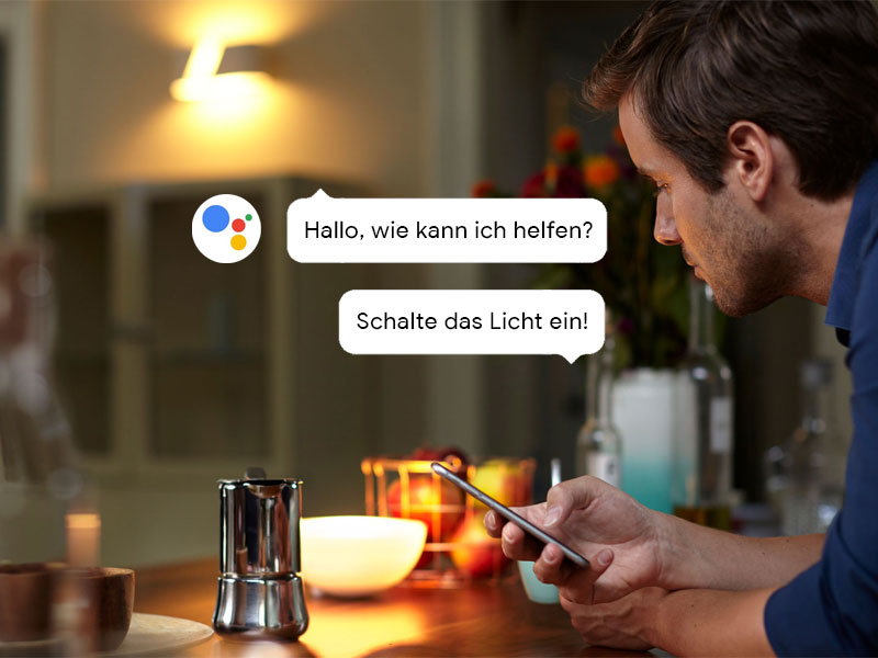 Junger Mann sitzt in der Küche mit Handy. Sprechbalse ok Google schalte das Licht ein. Die Lampen leuchten von Philips Hue